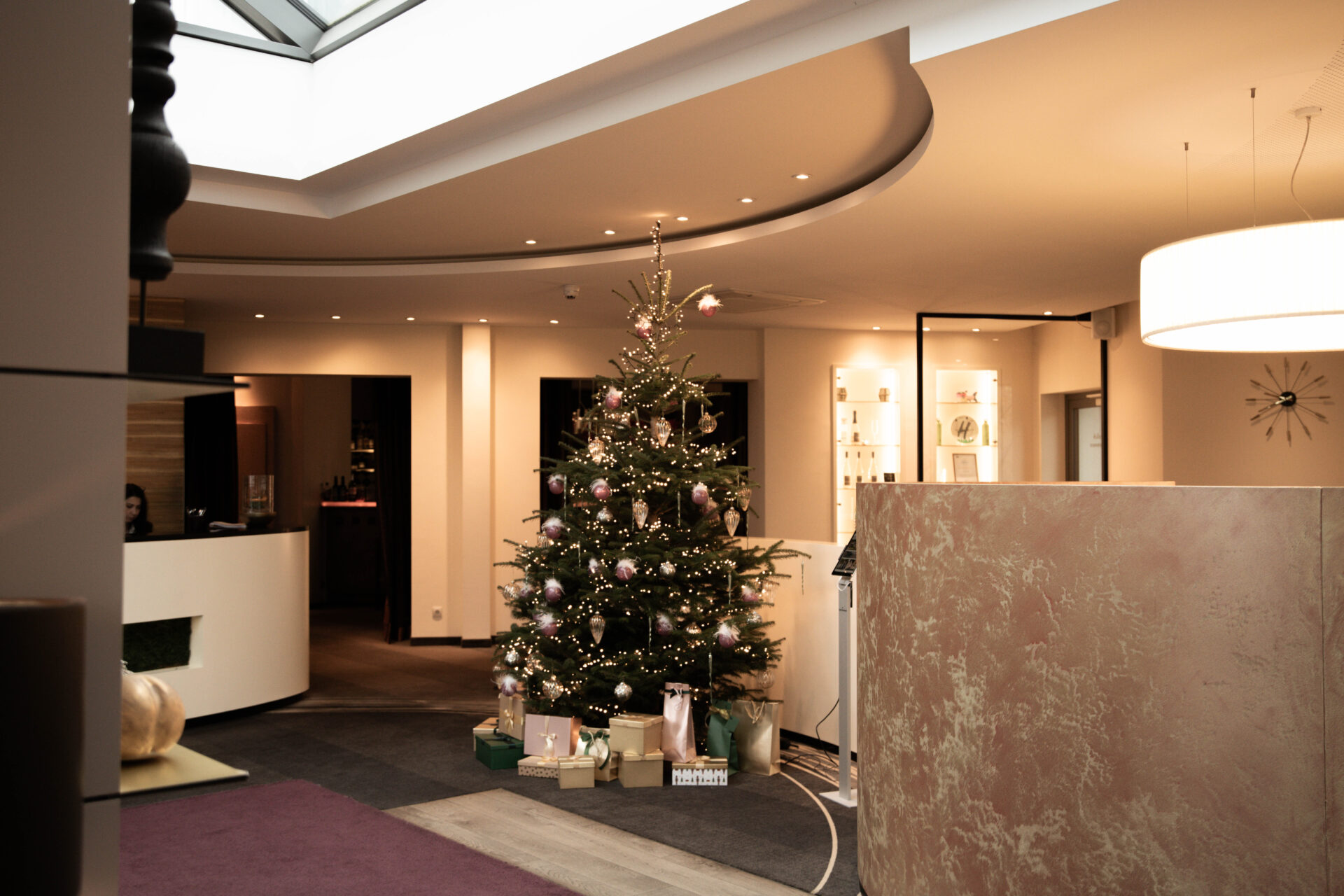 In einem modernen, sanft beleuchteten Lobbybereich steht ein geschmückter Weihnachtsbaum mit verpackten Geschenken darunter.