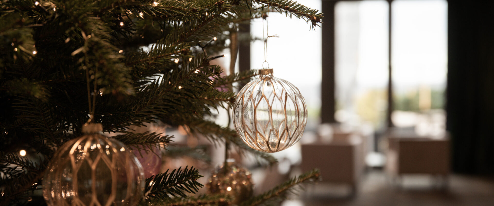 Nahaufnahme eines geschmückten Weihnachtsbaums mit Lichtern und Glasornamenten vor unscharfem Innenhintergrund.