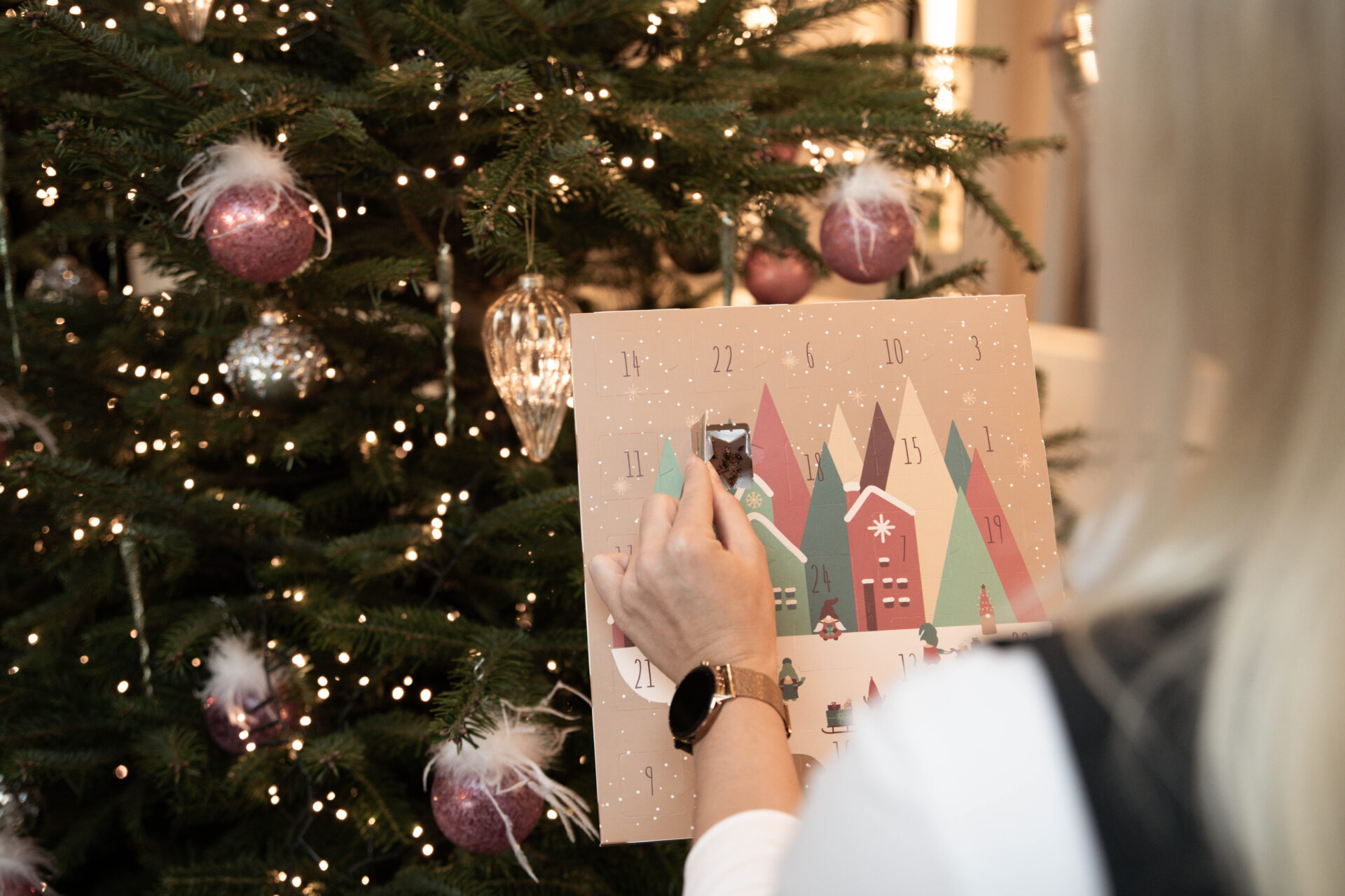 Eine Person öffnet ein Türchen an einem Adventskalender neben einem mit Lichtern und Ornamenten geschmückten Weihnachtsbaum.