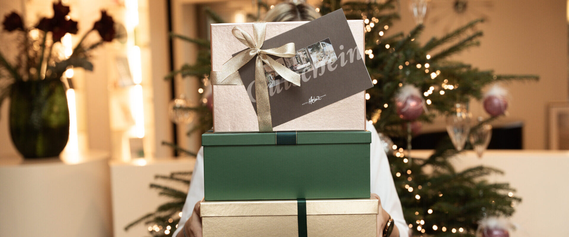 Eine Person hält drei verpackte Geschenke vor einem geschmückten Weihnachtsbaum mit Lichtern und Ornamenten.