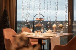 Ein gemütlicher, für zwei Personen gedeckter Tisch zum Tee mit Leckereien, Champagner und Lichterketten, die im Fenster dahinter funkeln.