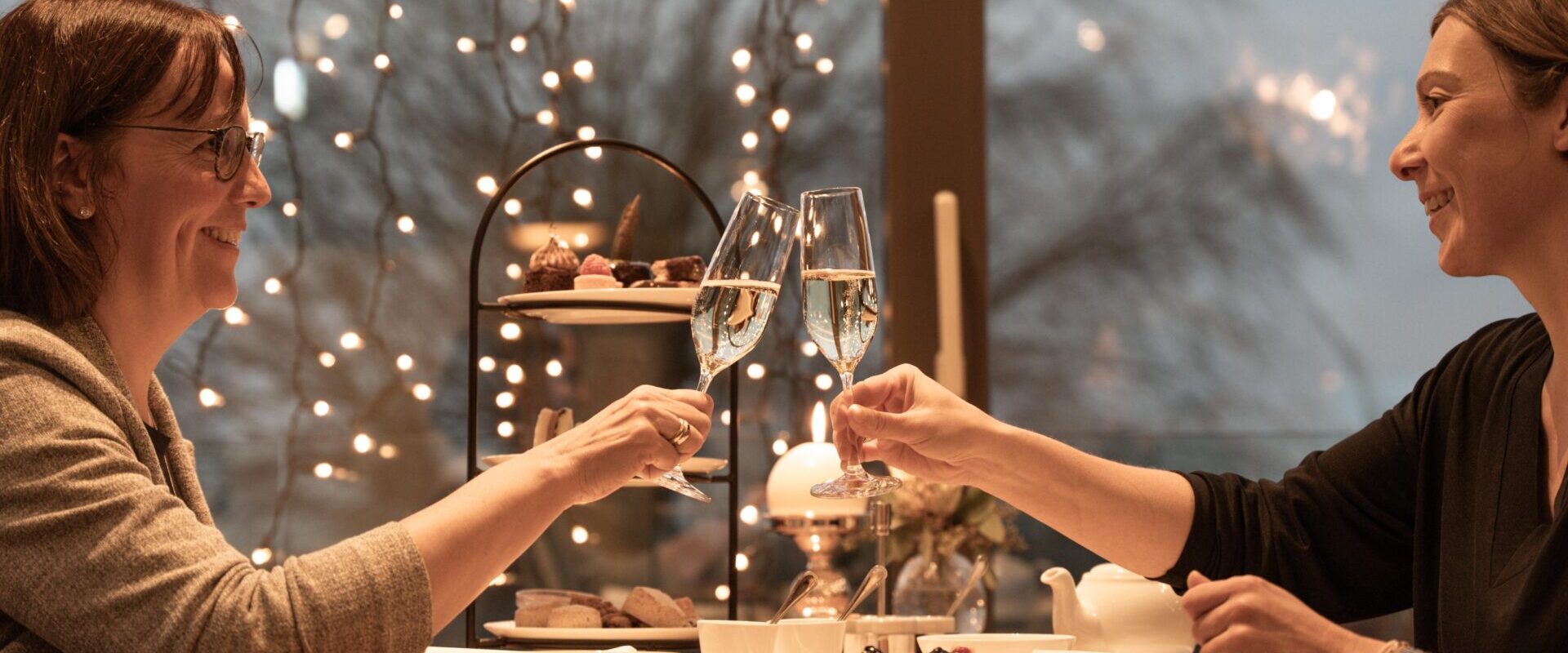 Zwei Frauen sitzen lächelnd an einem gedeckten Tisch und stoßen mit Champagnergläsern an, im Hintergrund funkeln Lichter.