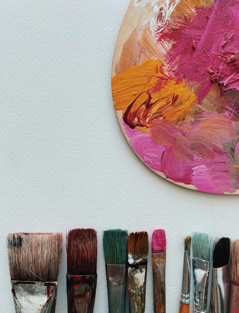 Eine Reihe gebrauchter Pinsel liegt unter einer bunten Künstlerpalette mit rosa, orangefarbener und gelber Farbe auf weißem Hintergrund.