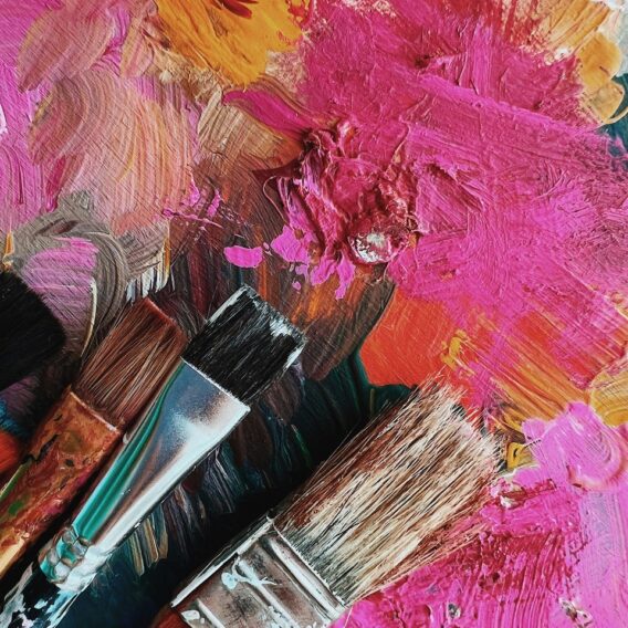Drei Pinsel liegen auf einer Palette, die mit dicker, bunter Acrylfarbe bedeckt ist, hauptsächlich in Rosa-, Orange- und Brauntönen.