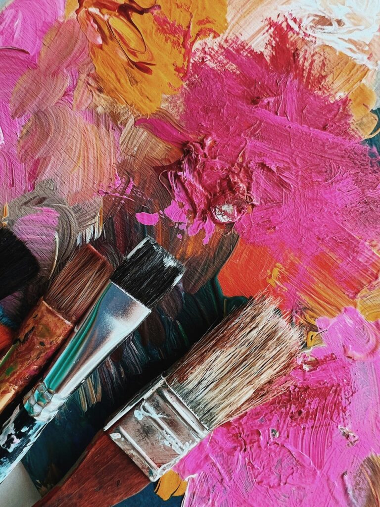 Drei Pinsel liegen auf einer Palette, die mit dicker, bunter Acrylfarbe bedeckt ist, hauptsächlich in Rosa-, Orange- und Brauntönen.