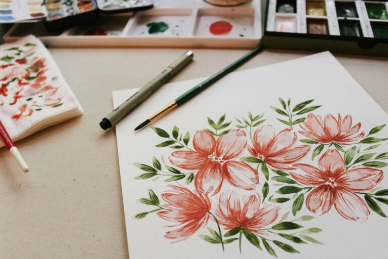 Ein Aquarell von rosa Blumen mit grünen Blättern auf Papier, umgeben von Pinseln, Stiften und Aquarellpaletten.