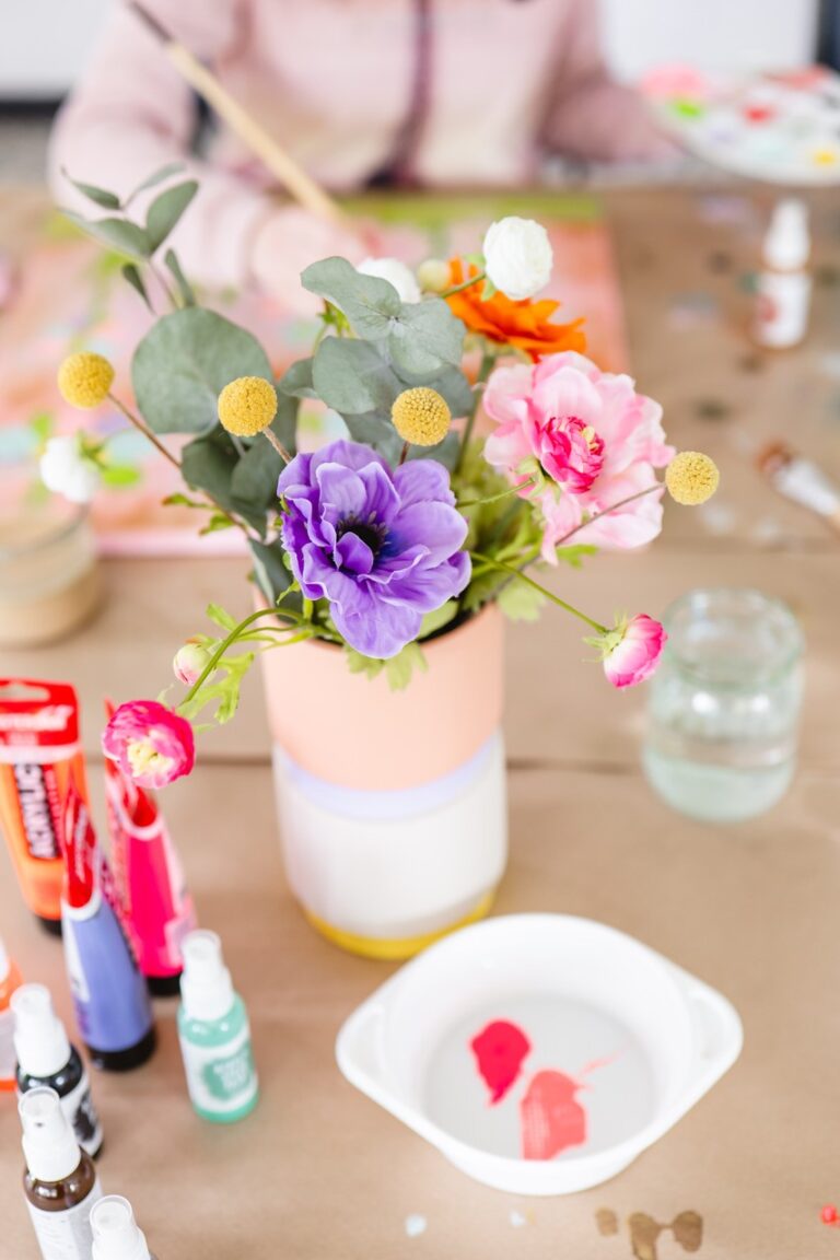 Auf einem Tisch steht ein bunter Blumenstrauß in einer Vase, im Hintergrund Farbtuben, Pinsel und eine malende Person.