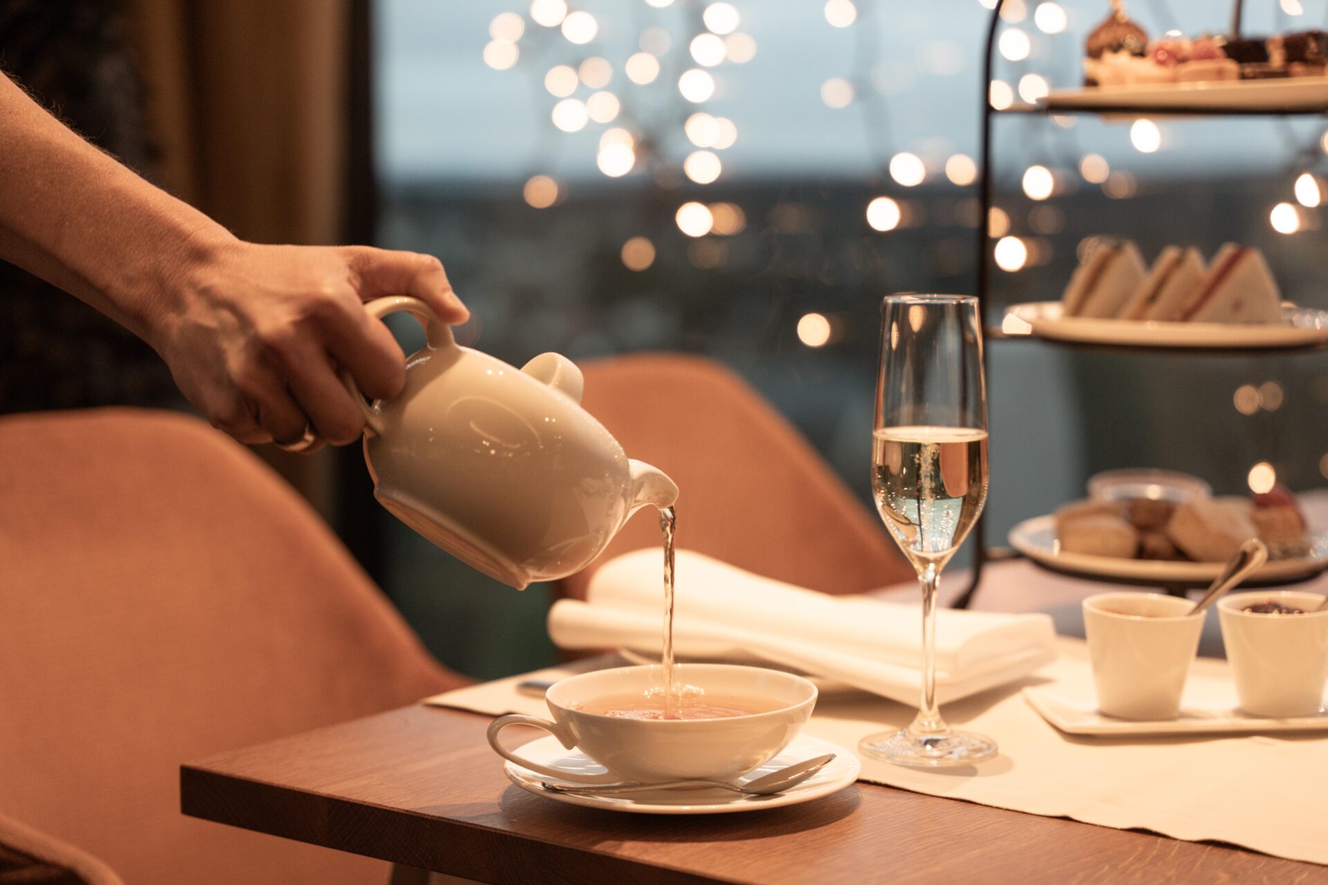 Eine Hand gießt Tee aus einer Teekanne in eine Tasse auf einem mit Gebäck, Champagner und Gewürzen gedeckten Tisch, im Hintergrund sanftes Licht.