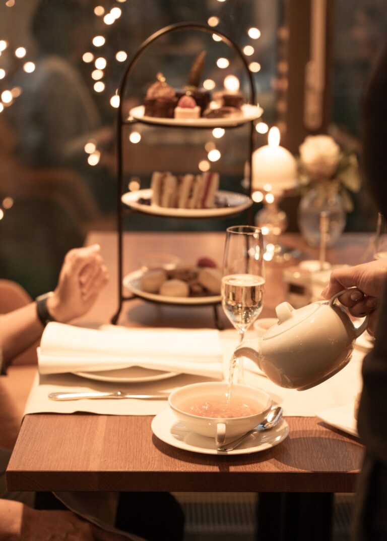 Eine Teekanne gießt Tee in eine Tasse auf einem für zwei Personen gedeckten Tisch mit Gebäck, Süßigkeiten und einem Glas Champagner bei warmer, sanfter Beleuchtung.