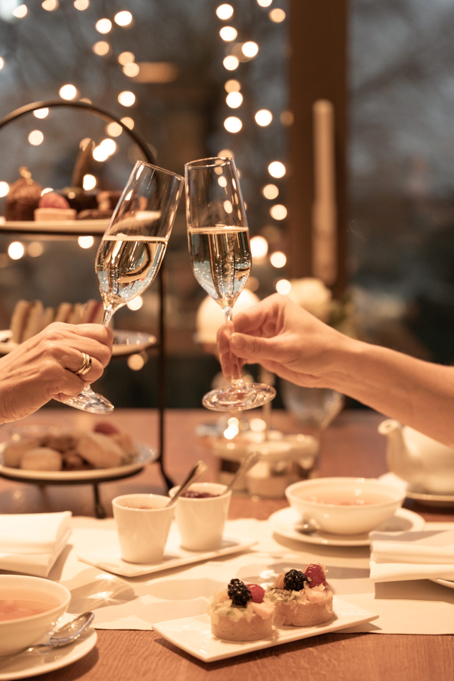Zwei Personen stoßen mit Champagnergläsern an einem mit Desserts, Tee und Gebäck gedeckten Tisch an, im Hintergrund schimmern warme Lichterketten.