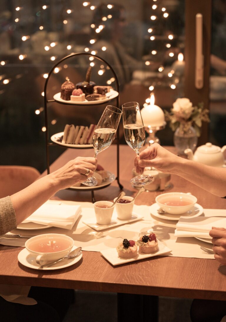 Zwei Personen stoßen mit Champagner an einem für Tee gedeckten Tisch an, im Hintergrund gibt es Gebäck, Desserts und funkelnde Lichter.