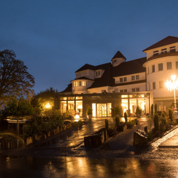 Großes, elegantes Hotel mit warmem Licht am Eingang, nassem Bürgersteig, der das Licht reflektiert, und einem dunklen Abendhimmel darüber.