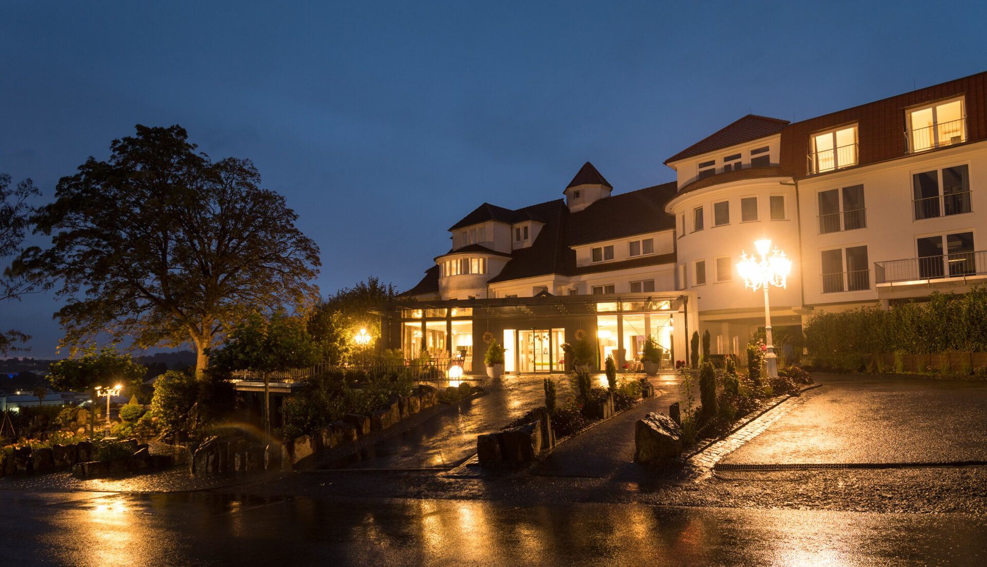 Großes, elegantes Hotel mit warmem Licht am Eingang, nassem Bürgersteig, der das Licht reflektiert, und einem dunklen Abendhimmel darüber.