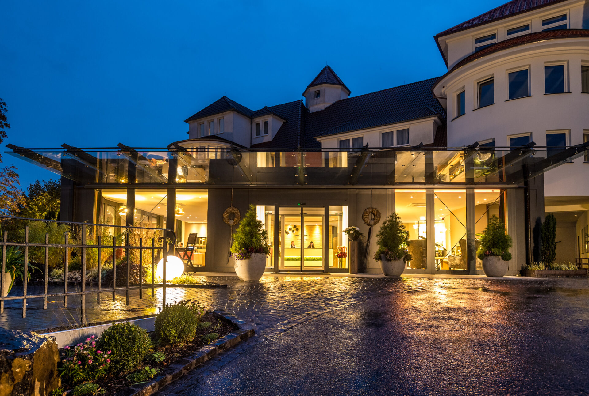 Hoteleingang in der Abenddämmerung, warm beleuchtet mit Glastüren, großen Topfpflanzen und einem nassen Steinweg, der das Licht reflektiert.