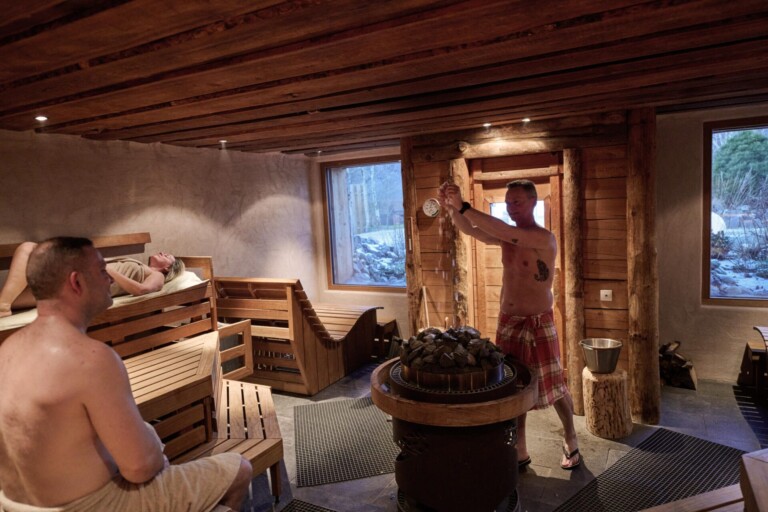 Ein Mann gießt Wasser auf heiße Saunasteine, während zwei andere auf Holzbänken in einem rustikalen Saunaraum mit großen Fenstern entspannen.