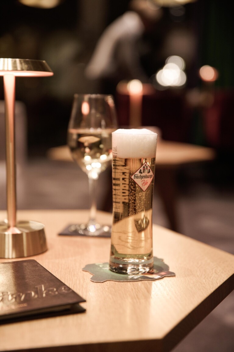 Ein Glas Bier und ein Glas Weißwein auf einem Tisch mit einer Lampe in einer schwach beleuchteten Bar oder einem Restaurant.