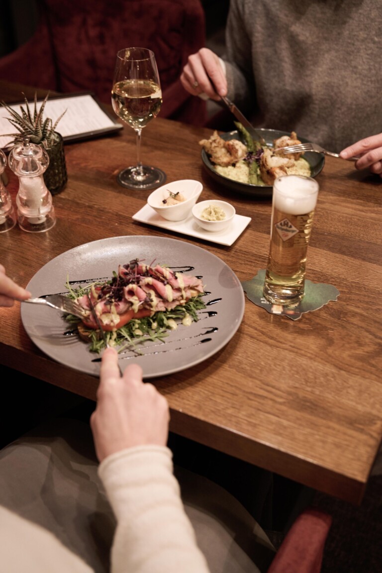 Zwei Personen essen in einem Restaurant mit Tellergerichten, einem Glas Weißwein und einem Glas Bier auf einem Holztisch.