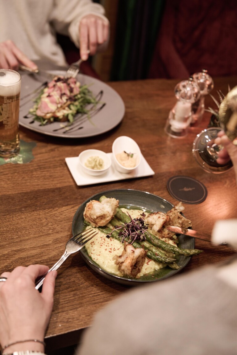 Zwei Personen essen in einem Restaurant auf einem Holztisch Teller mit gegrillten Garnelen, Spargel und Kartoffelpüree sowie Getränken und Gewürzen.