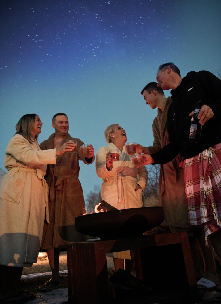 Fünf Personen in Bademänteln lachen am Feuer unter Sternenhimmel, halten Getränke, teils mit Decke um die Hüfte. Der Boden ist leicht verschneit.