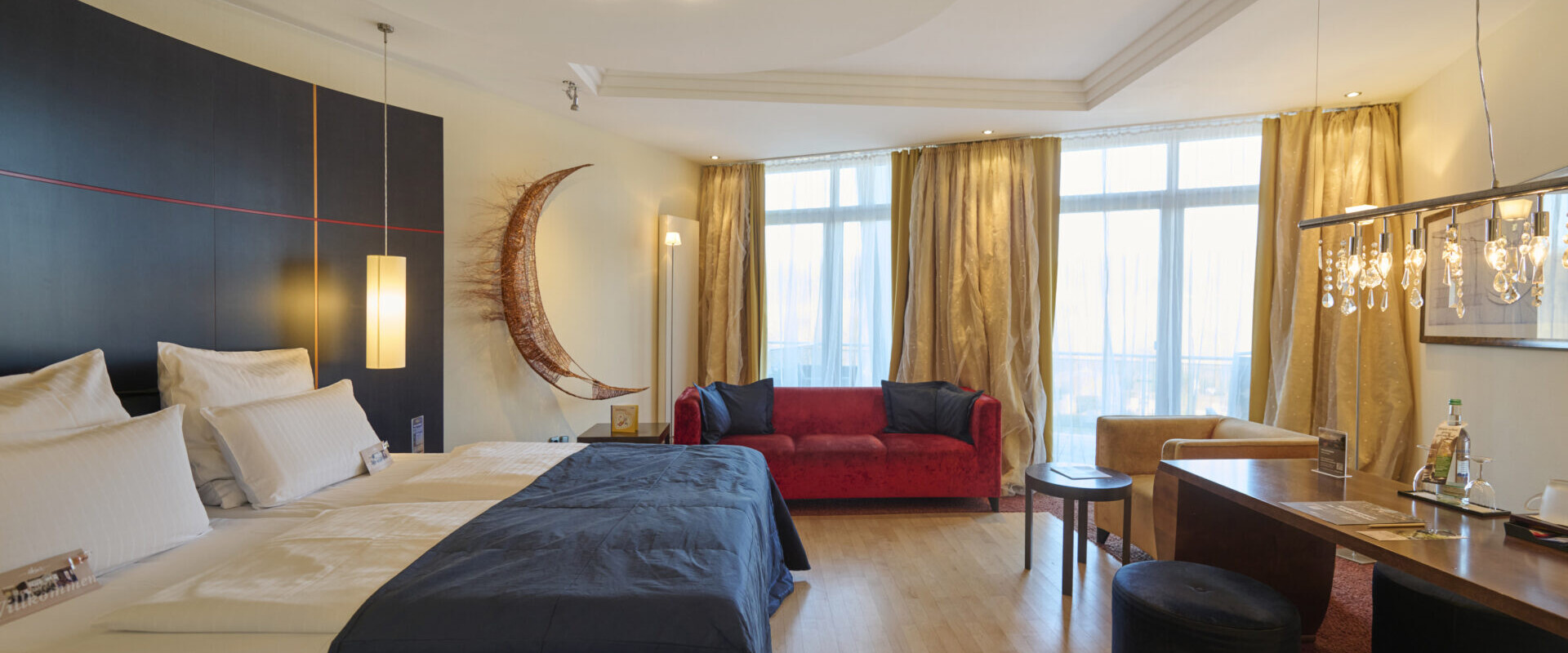 Geräumiges Hotelzimmer mit zwei Betten, einem roten Sofa, großen Fenstern, moderner Beleuchtung und einem Schreibtisch mit Teeservice.