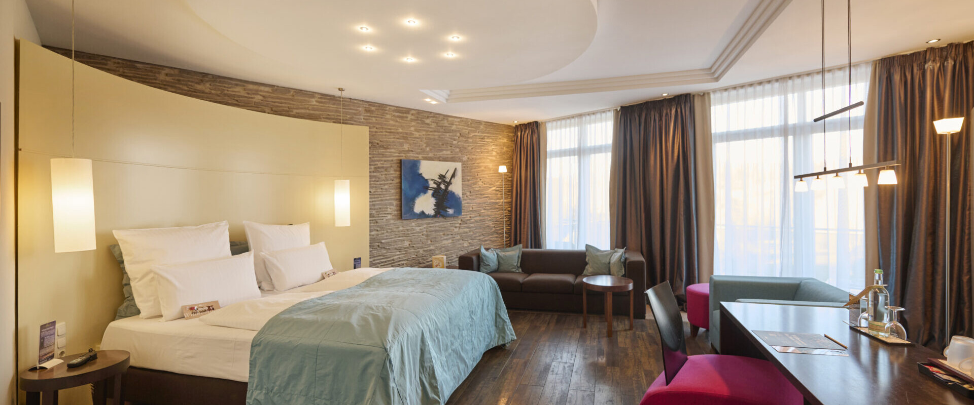 Modernes Hotelzimmer mit großem Bett, Sofa, Schreibtisch und raumhohen Fenstern mit dunklen Vorhängen, warm beleuchtet und stilvoll eingerichtet.