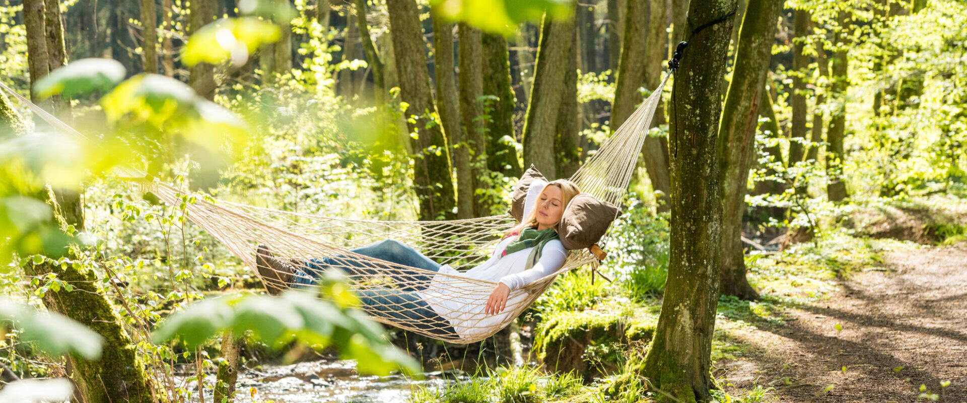 Zwei Menschen entspannen gemeinsam in einer zwischen Bäumen aufgespannten Hängematte in einem sonnenbeschienenen, grünen Wald in der Nähe eines kleinen Baches.