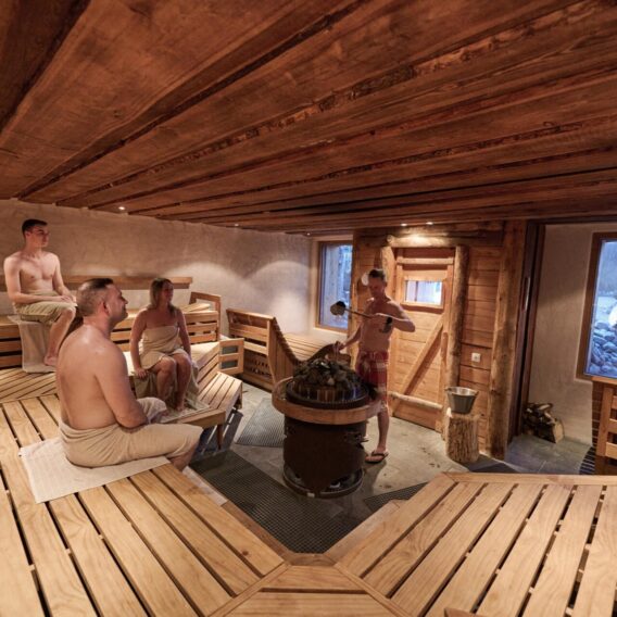 Four people wrapped in towels sit on wooden benches inside a spacious sauna with wooden walls and ceiling. One person pours water over hot stones while a window reveals a snowy outdoor scene.