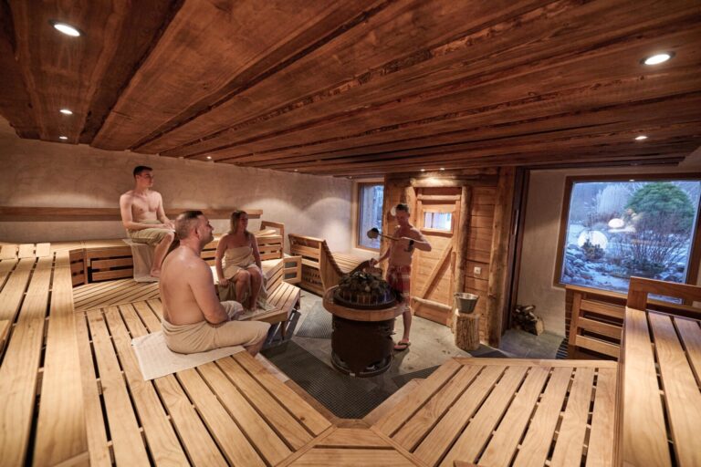 Four people wrapped in towels sit on wooden benches inside a spacious sauna with wooden walls and ceiling. One person pours water over hot stones while a window reveals a snowy outdoor scene.