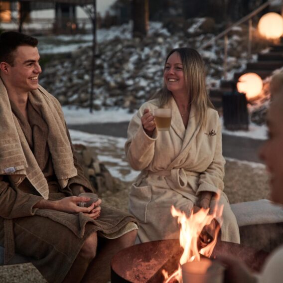 Three people in bathrobes sit around an outdoor fire pit, holding mugs and smiling. Snow is visible on the ground and large glowing lights are in the background, creating a cozy winter atmosphere.