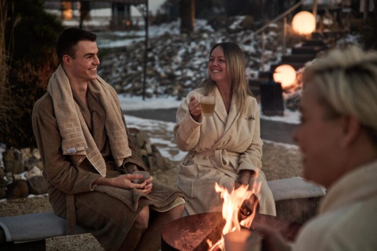 Drei Personen in Bademänteln genießen im Winter draußen an einer Feuerstelle heiße Getränke, lächeln und entspannen sich mit über die Schultern drapierten Handtüchern.