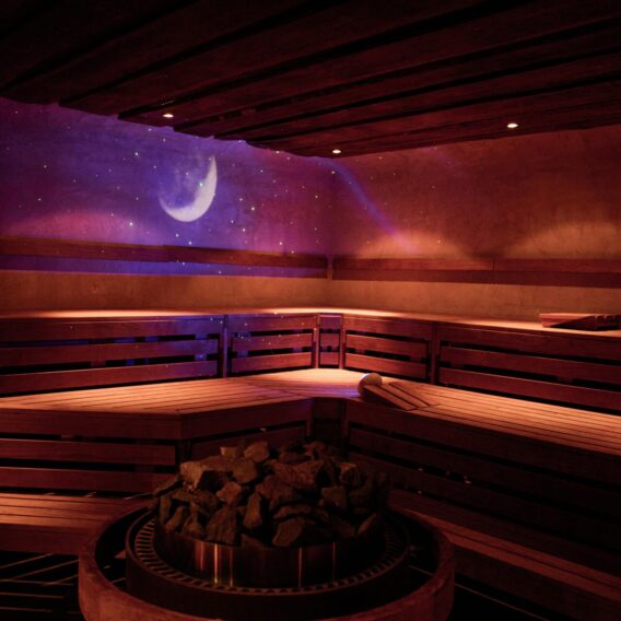 A dimly lit sauna with wooden benches, a pile of sauna stones in the center, and a crescent moon light projection on the wall, creating a calming, nighttime atmosphere.