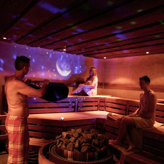 Three people relax in a dimly lit sauna with wooden benches; one person pours water on hot stones while two others sit, and purple lights with star and moon projections decorate the wall.