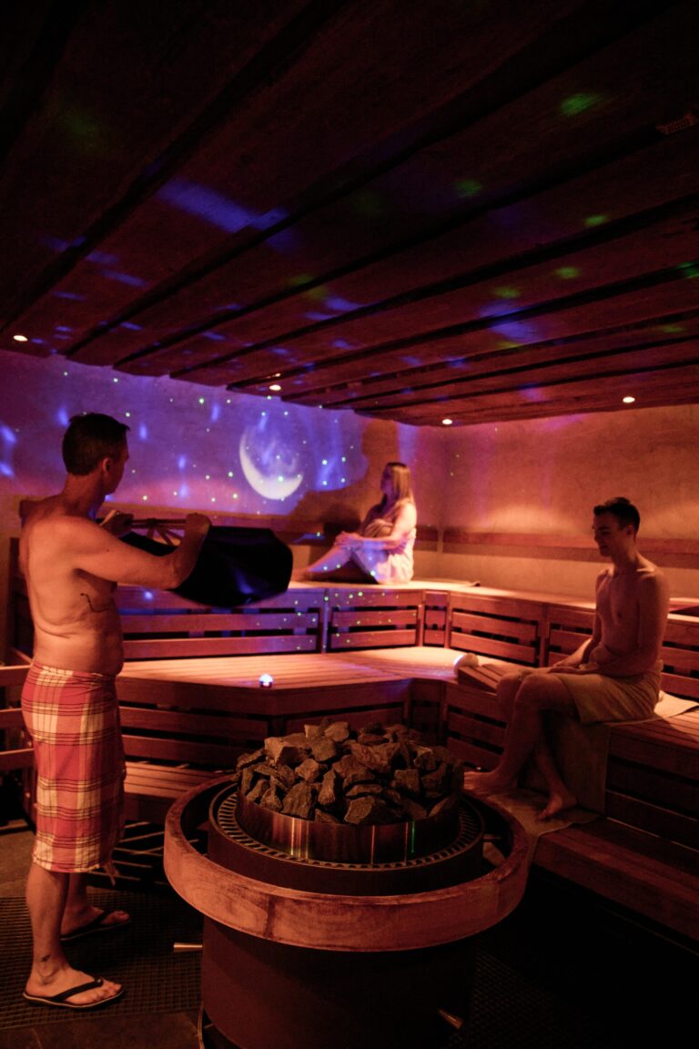 Drei Personen in Handtüchern entspannen sich in einer Holzsauna mit violetter Stimmungsbeleuchtung und einer Sternenprojektion an der Wand.