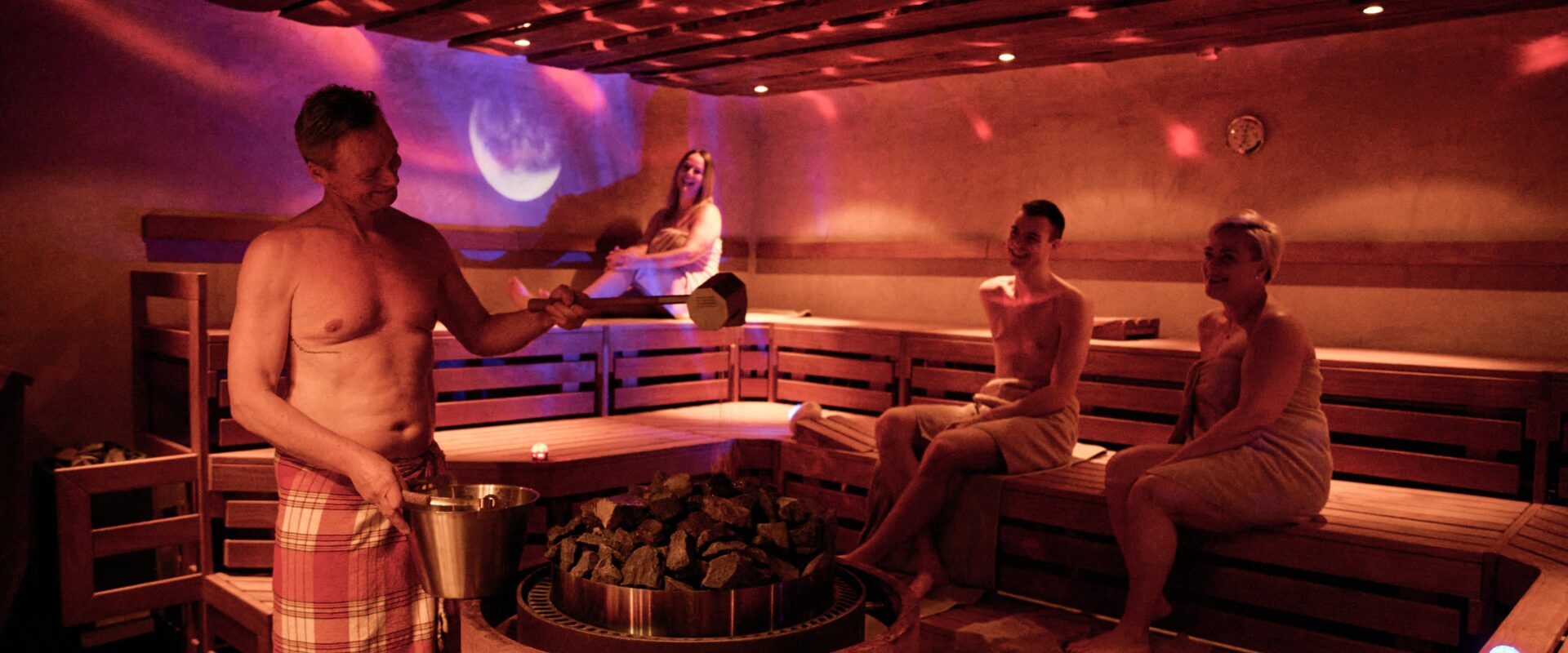 Vier Personen in Handtüchern entspannen sich in einer schwach beleuchteten Sauna, während eine Person Wasser über heiße Steine gießt, um Dampf zu erzeugen.