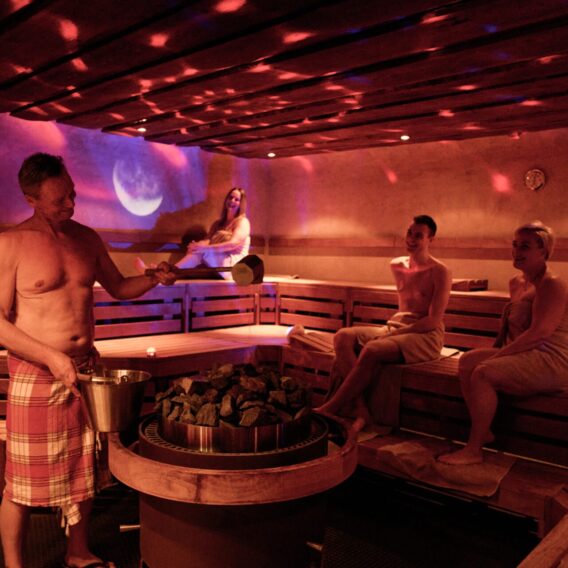 Four people are in a dimly lit sauna. One man wearing a towel pours water on hot stones, while three women, wrapped in towels, sit on wooden benches, relaxing and enjoying the steam.