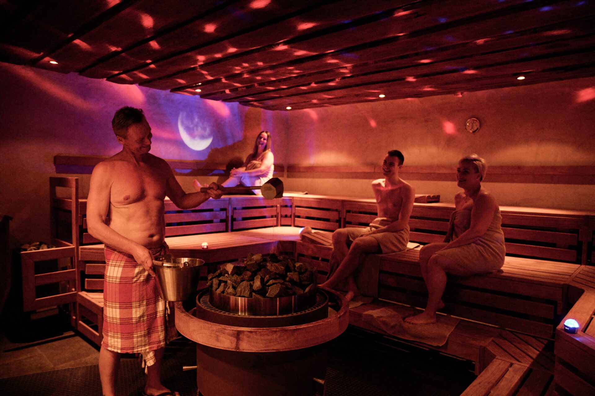 Four people are in a dimly lit sauna. One man wearing a towel pours water on hot stones, while three women, wrapped in towels, sit on wooden benches, relaxing and enjoying the steam.