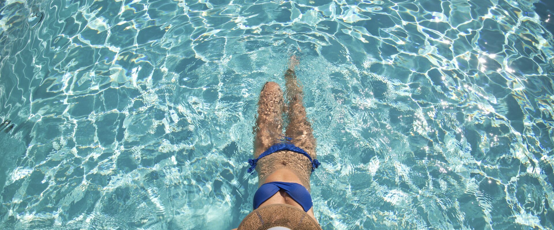 Der Außenpool: Eine Frau in einem blauen Bikini und einem breitkrempigen Hut entspannt sich am Rande eines Swimmingpools und hält ihre Beine ins Wasser.