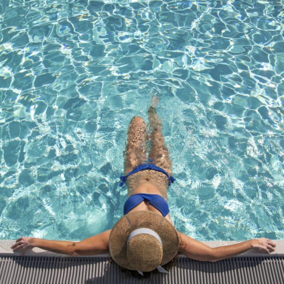 Der Außenpool: Eine Frau in einem blauen Bikini und einem breitkrempigen Hut entspannt sich am Rande eines Swimmingpools und hält ihre Beine ins Wasser.