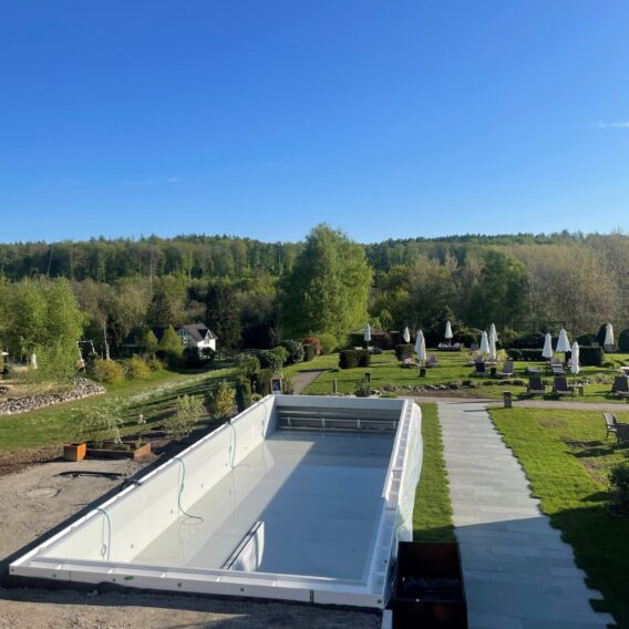 Ein rechteckiger, leerer Swimmingpool auf einer Terrasse überblickt einen grünen Garten mit Sonnenschirmen und Bäumen unter einem klaren blauen Himmel.