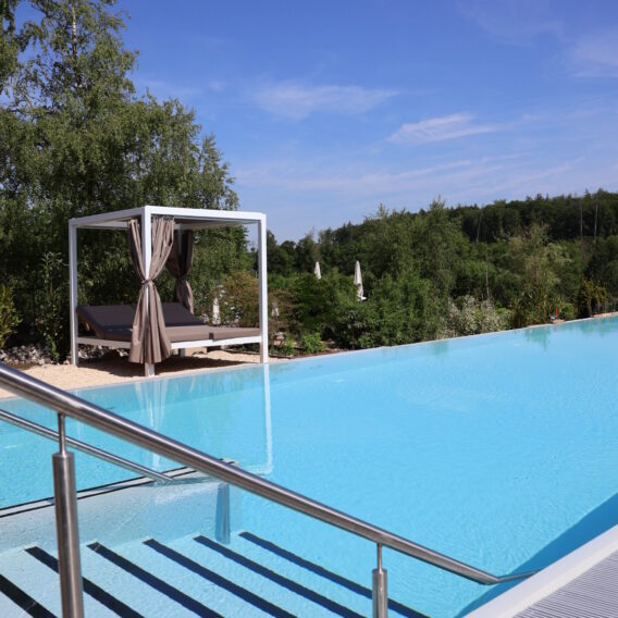 Der Außenpool: Infinity-Pool mit Metallgeländer, Himmelbett am Beckenrand und üppigem Grün unter einem strahlend blauen Himmel.