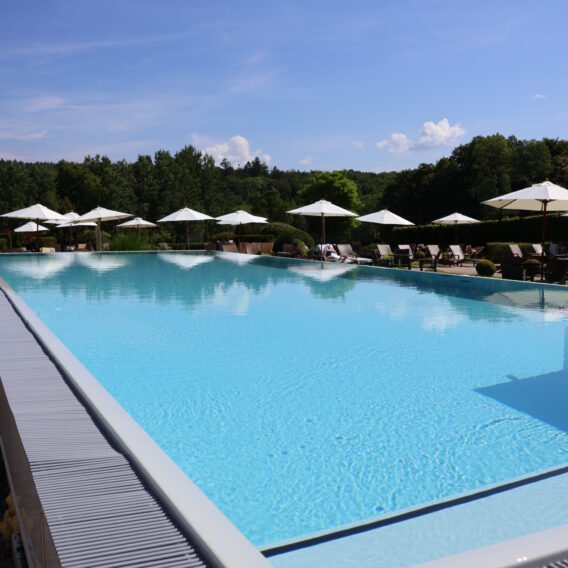 Ein Außenpool mit klarem, blauem Wasser, umgeben von Liegestühlen und weißen Sonnenschirmen, vor der Kulisse von Bäumen.