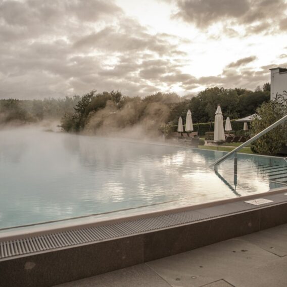 Der Außenpool: Infinity-Pool mit aufsteigendem Nebel, umgeben von Liegestühlen und Sonnenschirmen, mit bewölktem Himmel und Bäumen im Hintergrund.