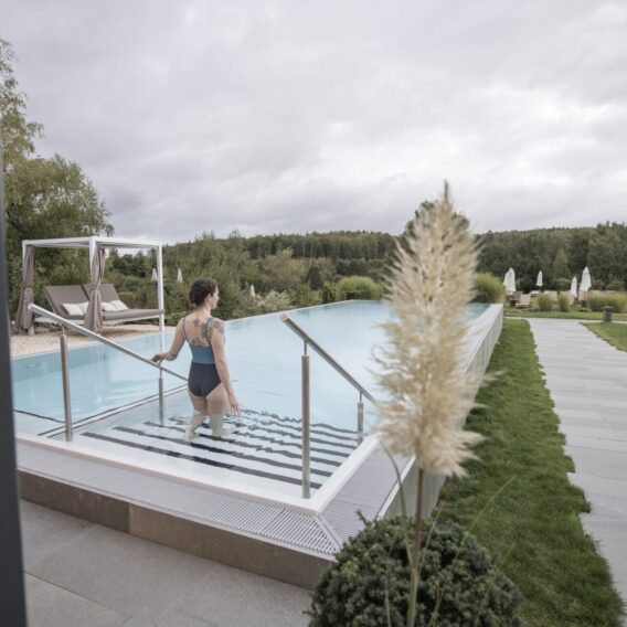 Der Außenpool: Eine Frau im Badeanzug steigt in ein Freibad, umgeben von Grünpflanzen, Liegestühlen und einem bedeckten Himmel.