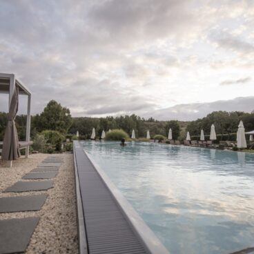 Der Außenpool: Infinity-Pool mit Liegestühlen und Sonnenschirmen, umgeben von Grün unter einem bewölkten Himmel bei Sonnenuntergang.