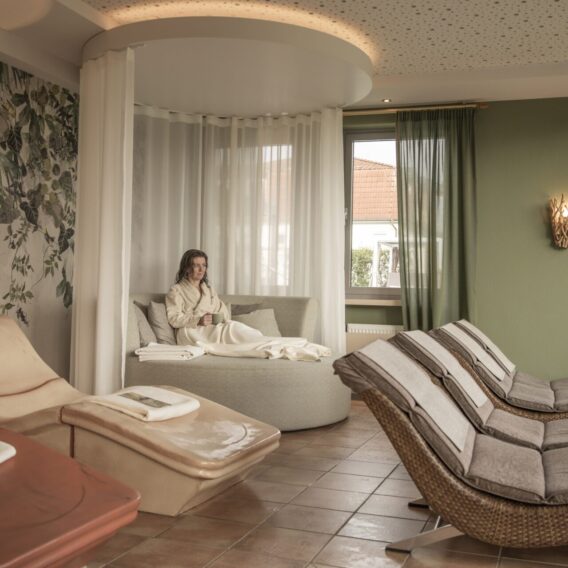 Ruhebereich der Therme: Eine Frau im Bademantel entspannt sich auf einem runden Sofa in einer gemütlichen Spa-Lounge mit Liegestühlen und sanfter Beleuchtung.