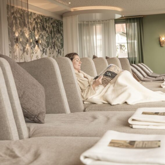 Ruhebereich der Therme: Eine Frau entspannt sich auf einem Liegestuhl in einem Wellnessbereich, zugedeckt mit einer Decke und einer Zeitschrift, während neben ihr weitere leere Liegen stehen.