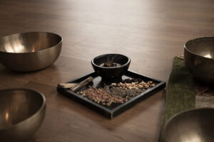 Räucher-Ritual: Ein Tablett mit getrockneten Kräutern, einer kleinen Schale und Weihrauch steht auf einem Holzboden, umgeben von großen Metallklangschalen.