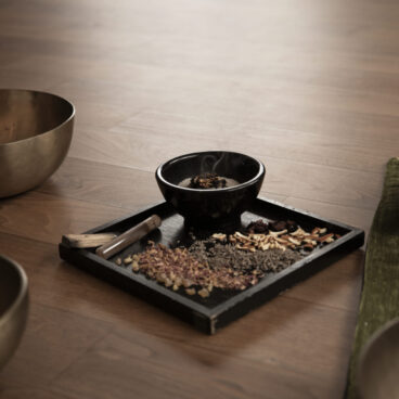 Räucher-Ritual: Ein Tablett mit getrockneten Kräutern, einer kleinen Schale und Weihrauch steht auf einem Holzboden, umgeben von großen Metallklangschalen.
