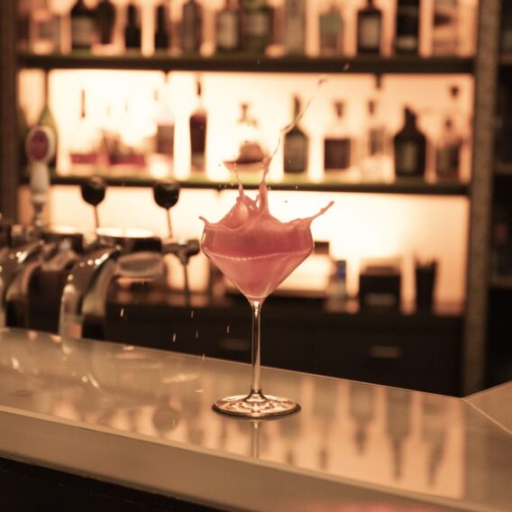Die Bar: Ein rosafarbener Cocktail plätschert aus einem Glas auf einem Bartresen, mit Flaschen und warmer Beleuchtung im Hintergrund.