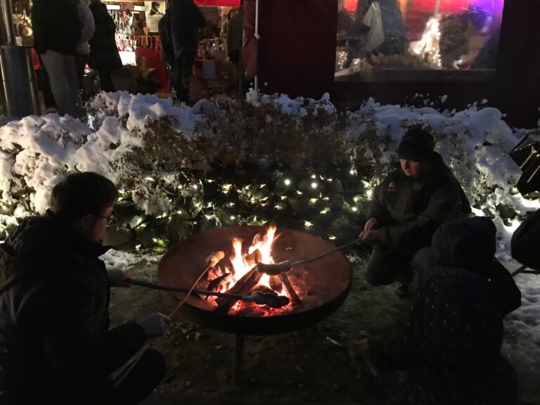 Adventsmarkt im Flürchen: Drei Personen braten nachts im Freien über einer offenen Feuerstelle, umgeben von Schnee und Lichterketten, mit einem festlichen Markt im Hintergrund.